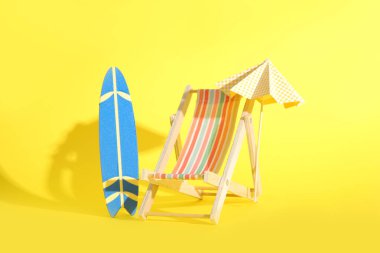 Sarı arka planda minyatür sörf tahtası, şezlonga ve şemsiyeye sahip yaratıcı kompozisyon