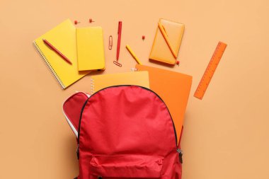 Kırmızı okul sırt çantası, turuncu arka planda defter, kalem ve cetvel.