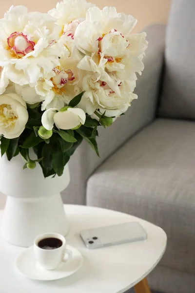 ソファの近くのテーブルの上にコーヒーと携帯電話のカップと白い牡丹の花瓶 — ストック写真