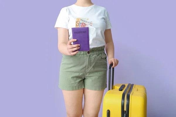 Young woman with suitcase, passport, ticket and money on lilac background. Travel concept