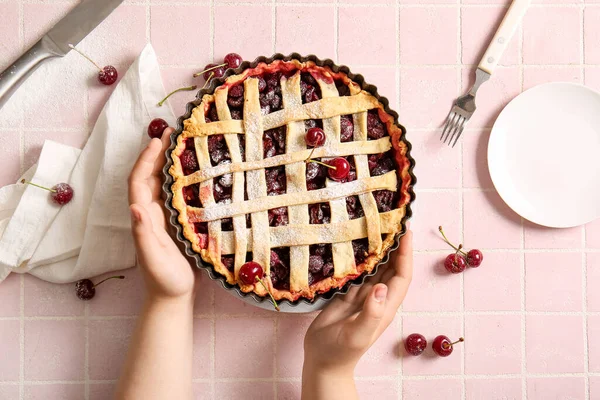 stock image Female hands holding baking dish with tasty cherry pie on pink tile background