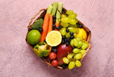 Pembe arka planda farklı taze meyve ve sebzelerle dolu hasır sepet.