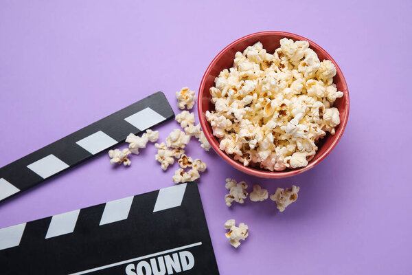 Bowl with tasty popcorn and clapperboard on purple background