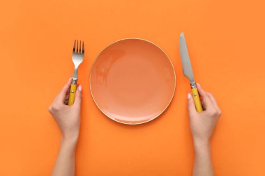 Çatal bıçaklı kadın elleri ve turuncu arka planda boş tabak.