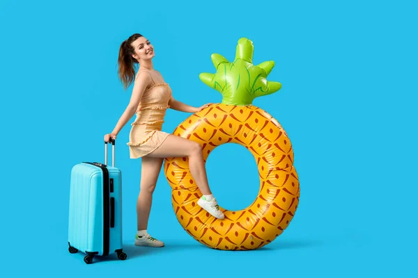 Young woman with suitcase and swim ring in shape of pineapple on blue background