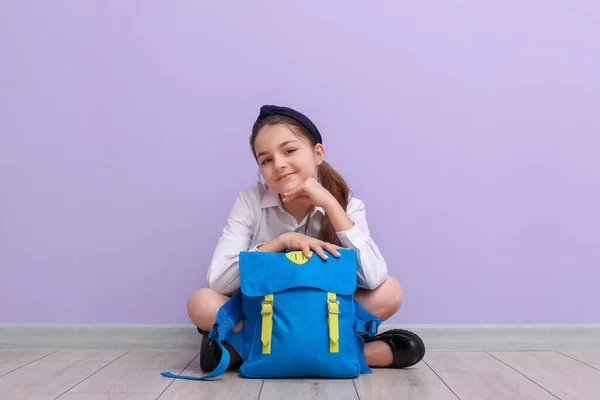 Kleines Schulmädchen Mit Rucksack Sitzt Neben Fliederfarbener Wand — Stockfoto