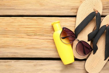 Terlikli güneş gözlüğü, hasır şapka ve ahşap arka planda bir şişe güneş kremi.