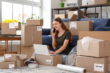 Paketli ve dizüstü bilgisayarı olan genç bir kadın evde cep telefonuyla konuşuyor.