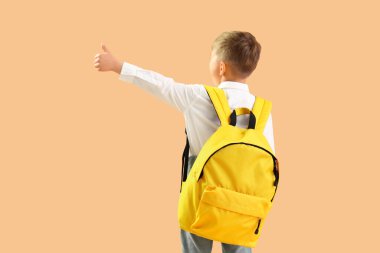 Sarı sırt çantalı küçük okul çocuğu turuncu arka planda baş parmak hareketi gösteriyor.