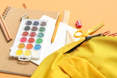 Turuncu arka planda boya ve kırtasiye malzemesi olan okul çantası, yakın plan.