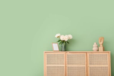 Yeşil duvarın yanındaki şifoniyerde fotoğraf çerçevesi ve biblo olan beyaz şakayıklar vazosu.