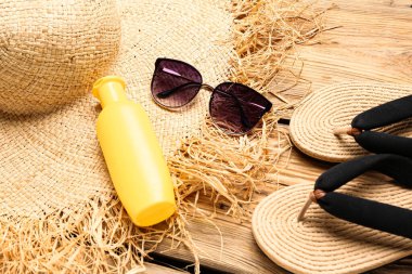 Terlikli güneş gözlüğü, hasır şapka ve kahverengi ahşap arka planda bir şişe güneş kremi.