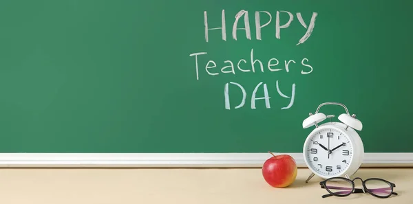 Text HAPPY TEACHERS DAY on green chalkboard, alarm clock, apple and eyeglasses on desk in classroom. Banner for design
