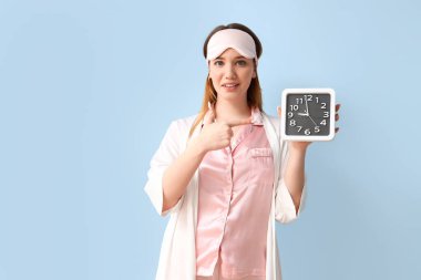 Pijamalı genç bir kadın mavi arka plandaki saate bakıyor.