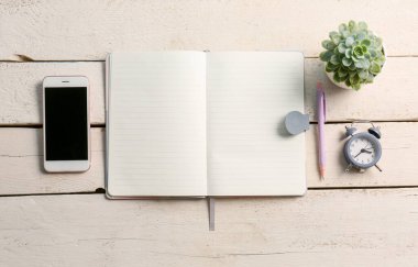 Beyaz ahşap arka planda boş bir defter, kalem, cep telefonu ve alarm saati