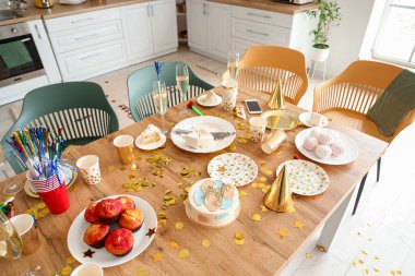 Mutfakta doğum günü pastası, ikram, şampanya ve parti şapkası olan yemek masası.