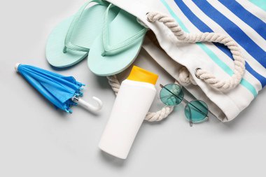 Güneş gözlüklü çanta, parmak arası terlik, mini şemsiye ve gri arka planda bir şişe güneş kremi.