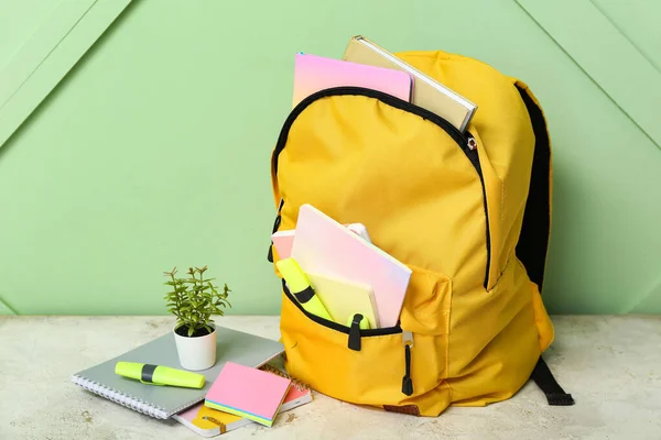 靠近绿墙的浅绿色桌子上有带笔记本 家居用品和标识的黄色校服背包 — 图库照片