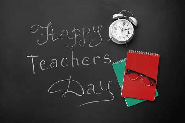Notebooks, alarm clock and text HAPPY TEACHERS DAY on black chalkboard