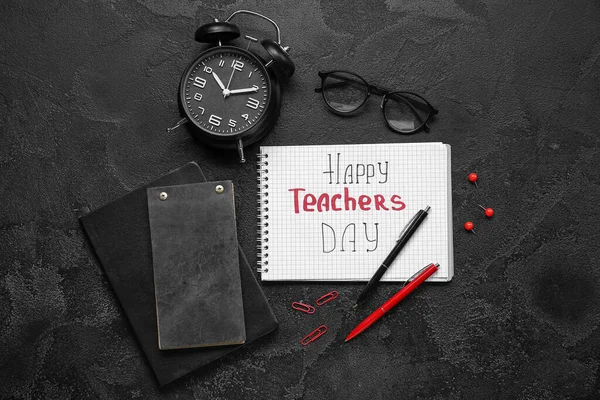 Notebooks with text HAPPY TEACHERS DAY, eyeglasses and alarm clock on grunge black background