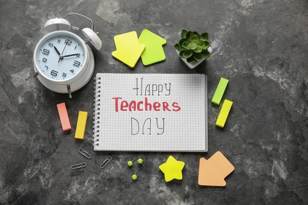 Notebook with text HAPPY TEACHERS DAY, alarm clock and different stationery on grunge black background