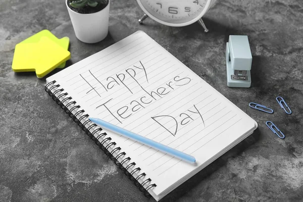 Notebook with text HAPPY TEACHERS DAY, alarm clock and different stationery on grunge black background