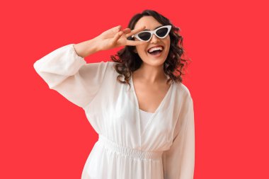 Güneş gözlüklü genç kadın kırmızı arka planda zafer işareti gösteriyor.