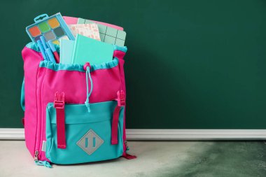 Üzerinde not defterleri, suluboya boyalar ve kalemler olan renkli okul çantası yeşil karatahtanın yanında.