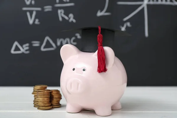 卒業キャップとブラックボードに対する白いテーブルの上にコインのスタックを持つ貯金箱 学生ローンの概念 — ストック写真