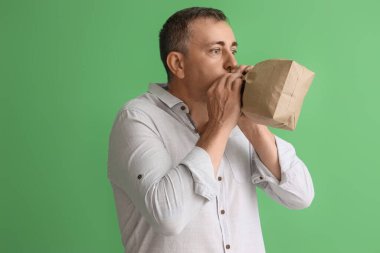 Yeşil arka planda panik atak geçiren, kağıt çantalı olgun bir adam.