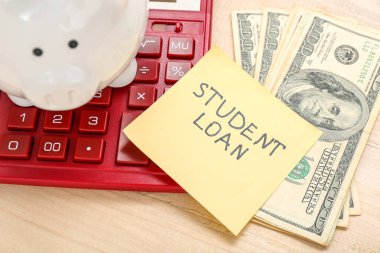 Tahta arka planda SÜDÜS kredisi, domuz kumbarası, hesap makinesi ve dolar banknotları olan yapışkan bir not.