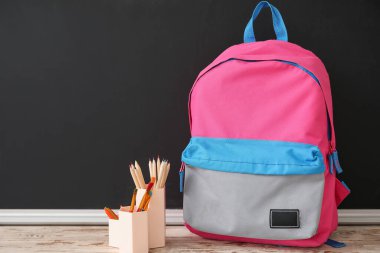 Siyah karatahtanın yanındaki ahşap masada bir kase kalem ve fırçayla pembe okul çantası.