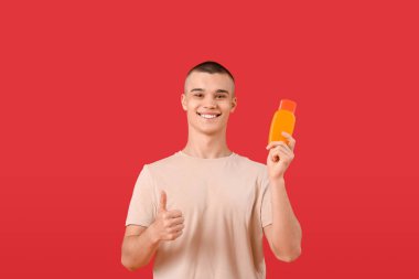 Güneş kremi şişesiyle mutlu genç adam kırmızı arka planda baş parmak hareketi gösteriyor.