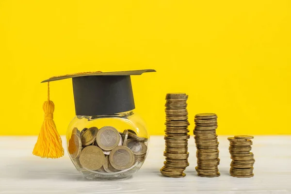 黄色の背景に木製のテーブルの上にお金 卒業キャップとコインのスタックを持つジャー 学生ローンの概念 — ストック写真
