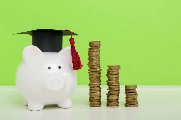 卒業キャップと緑の背景に白いテーブルの上にコインのスタックを持つ貯金箱 学生ローンの概念 — ストック写真