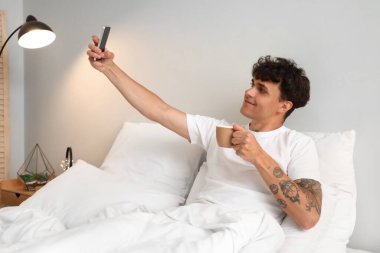 Bir fincan kahve ve cep telefonuyla yatakta selfie çeken genç bir adam.