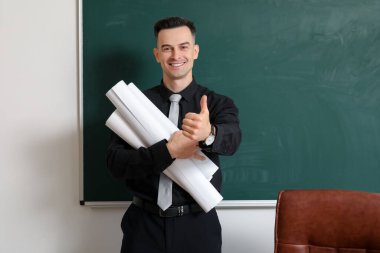 Sınıfta yazı tahtasının yanında kağıt tomarları olan erkek öğretmen.