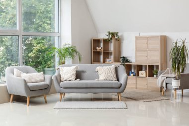 Aydınlık oturma odasının içinde şık gri kanepe, koltuk, raf ünitesi ve ev bitkileri var.