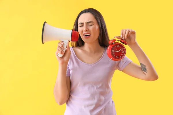 Stressed young woman with alarm clock shouting into megaphone on yellow background. Deadline concept