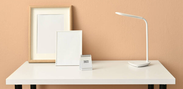 Stylish workplace with lamp, blank photo frames and calendar near beige wall. Banner for design