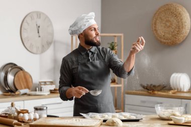 Male baker sprinkling raw buns with flour at table in kitchen