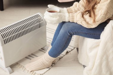 Elinde bir fincan sıcak kahve olan kadın radyatörün yanında ısınıyor.