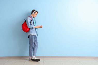 Sırt çantalı ve cep telefonlu bir erkek öğrenci mavi duvarın yanında kulaklıkla müzik dinliyor.