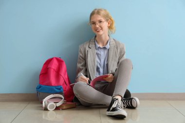Sırt çantalı ve kulaklıklı mutlu bayan öğrenci mavi duvarın yanında oturuyor.