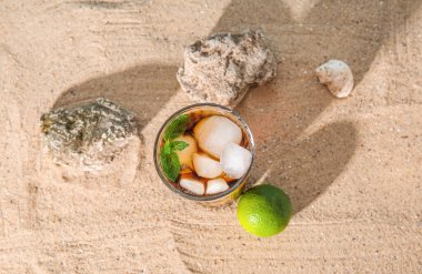 Bir bardak soğuk Küba Libre kokteyli ve kumdaki taşlar.