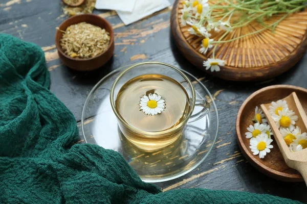 stock image Cup of hot tea and bowl with dried chamomile on black wooden background