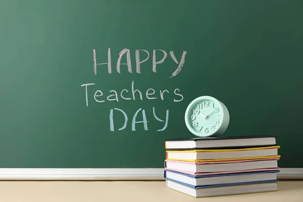 Text HAPPY TEACHERS DAY on green chalkboard with alarm clock and books in classroom