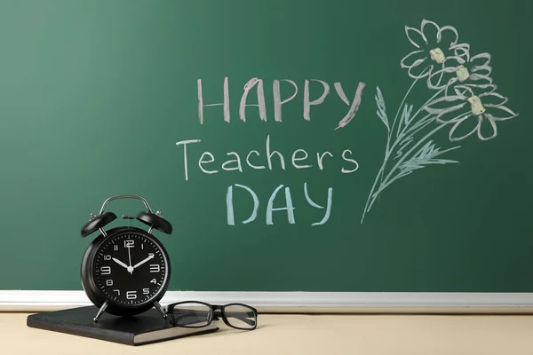 Drawn flowers and text HAPPY TEACHERS DAY on green chalkboard with alarm clock in classroom