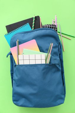 Yeşil arka planda not defterleri ve kalemlerle dolu mavi okul çantası.