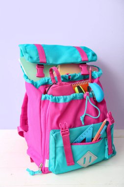 Renkli okul çantası, içinde not defterleri, kalem kutusu ve zımba var. Beyaz ahşap masa üzerinde, leylak duvarın yanında.